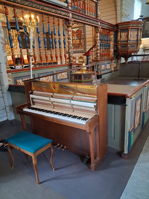 Oppreist piano med front- og toppanel tatt av, i en rikt dekorert trekirke, med pianokrakk foran